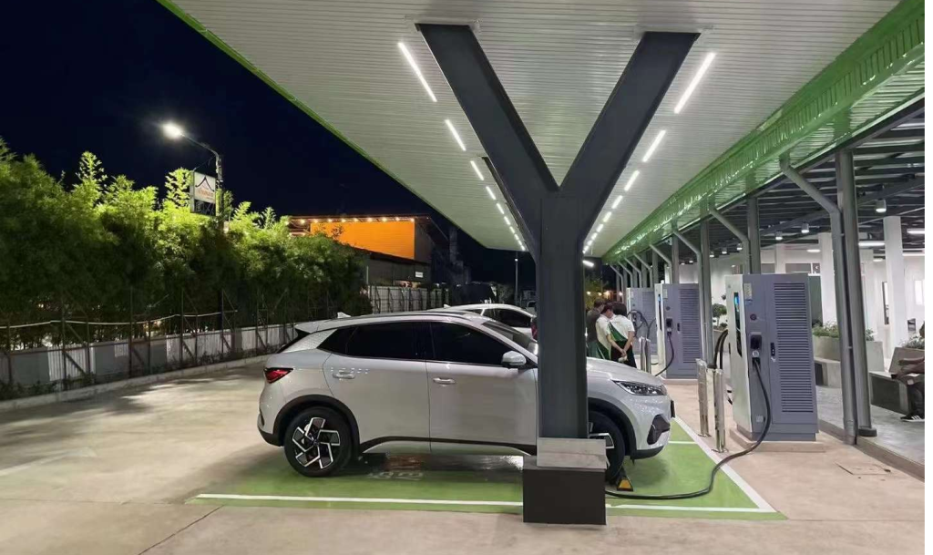 A night view of an electric vehicle charging station with overhead lighting, multiple charging units, a white SUV plugged in, and people in the background.