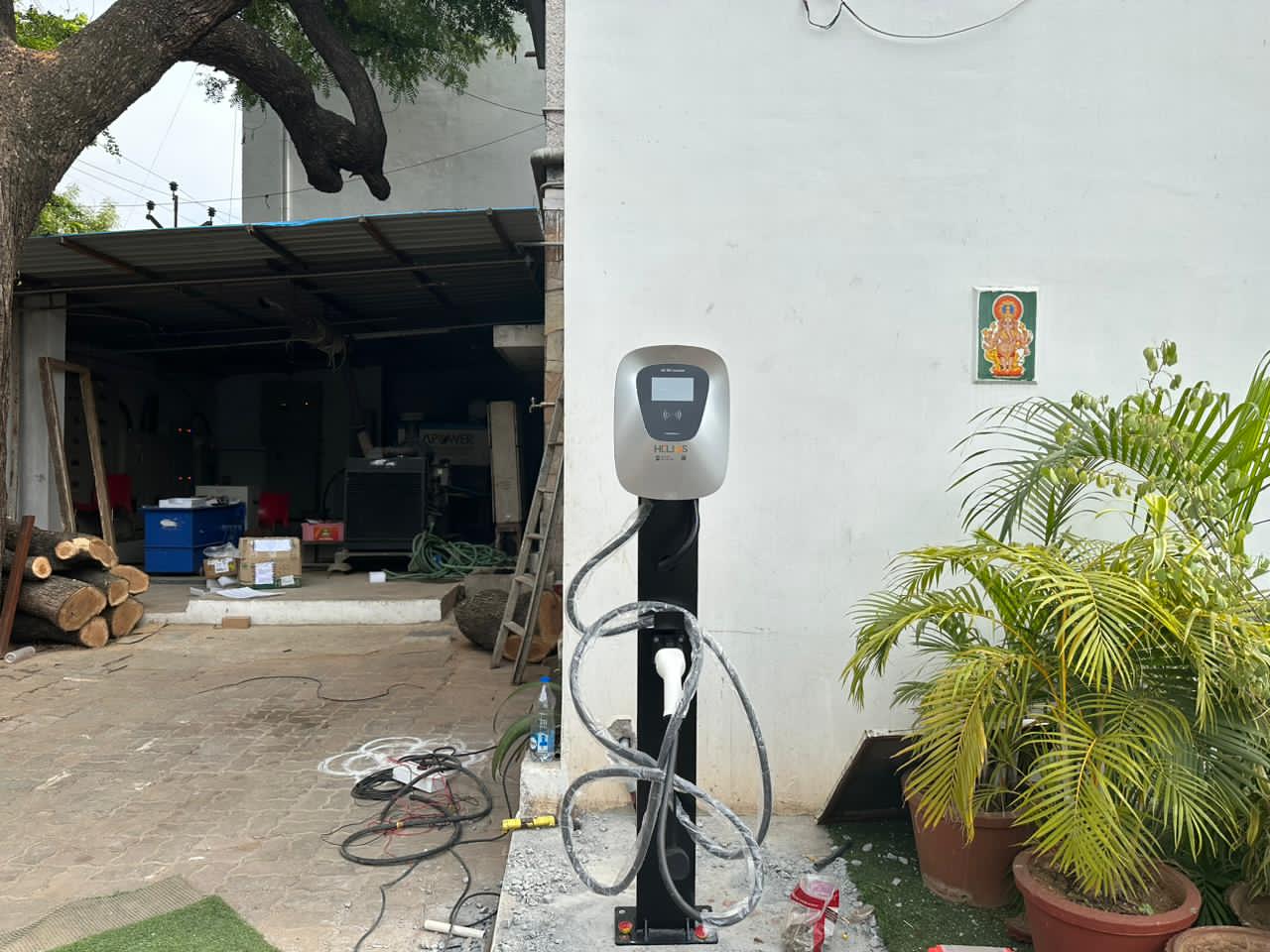 Electric vehicle charging station set up outdoors next to a white wall with religious iconography and green potted plants.